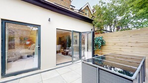 A modern patio area with a glass door leading to a living room, a glass-top table, and a wicker bench. - Prime 3BR Flat With Private Patio-Walk to Tube (Richmond)