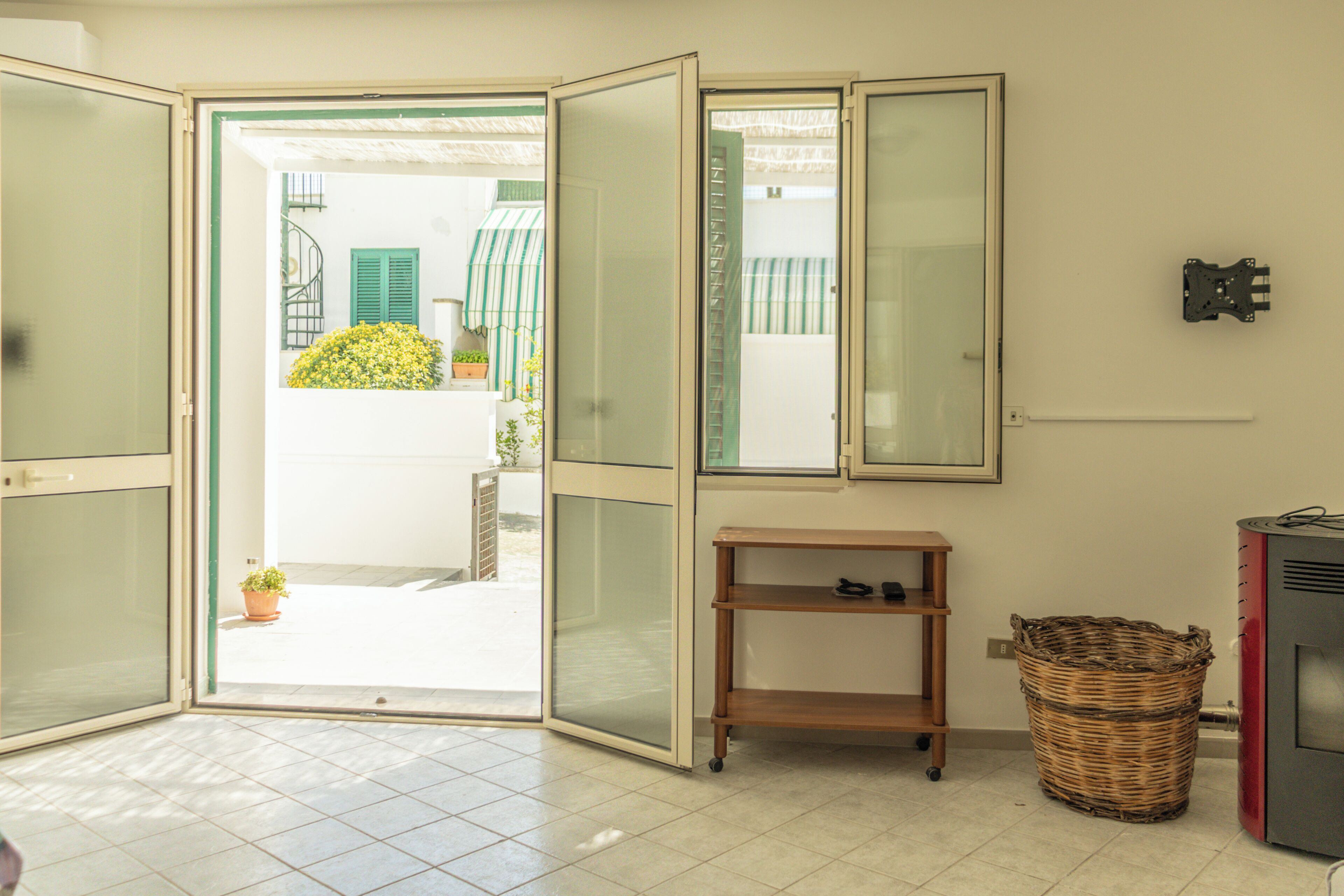 Habitació amb una porta corredissa de vidre que dona a un balcó, una petita estant de fusta, una cistella de vimbles i un aparell vermell.