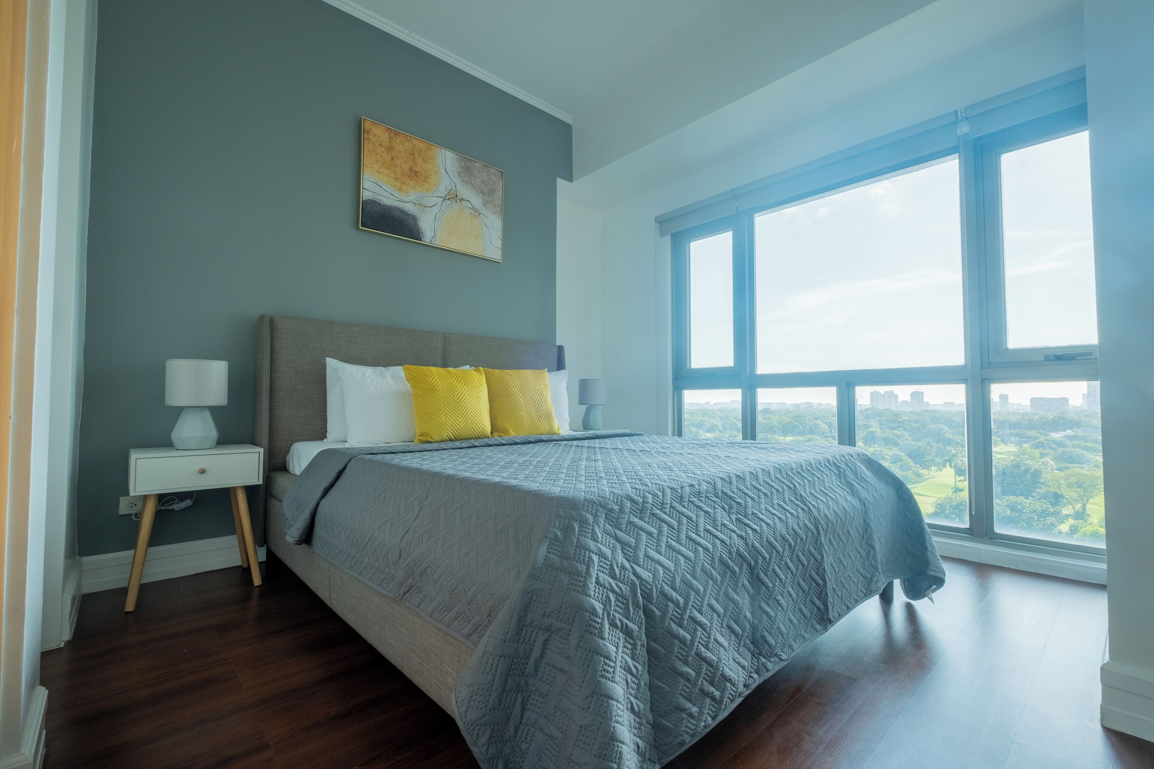A bedroom with a large window, a bed with a grey quilt, a nightstand with a lamp, and a painting on the wall.