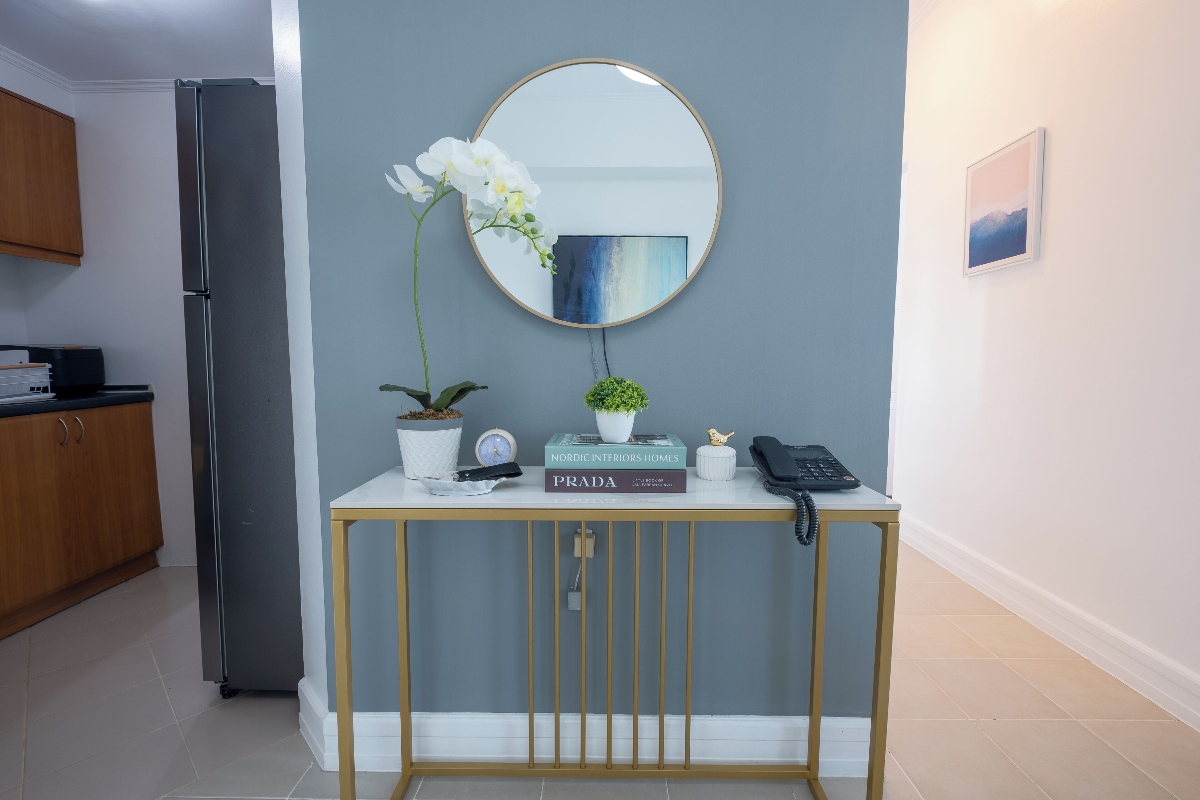 A modern kitchen with a stainless steel refrigerator, a wooden cabinet, and a console table with a mirror, books, a potted plant, and a phone.