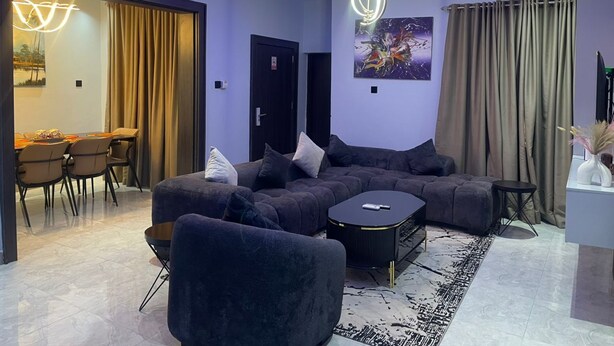 A modern living room with a sectional sofa, a coffee table, and a dining area with chairs and a table. - The Address 127 - Refined Apartments (Abuja)