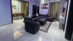 A modern living room with a sectional sofa, a round coffee table, and a dining area with chairs. - The Address 127 - Refined Apartments (Abuja)