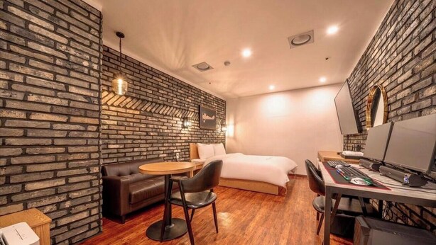Una habitación con pared de ladrillo, una cama, un escritorio con computadora, una silla y una pequeña mesa.