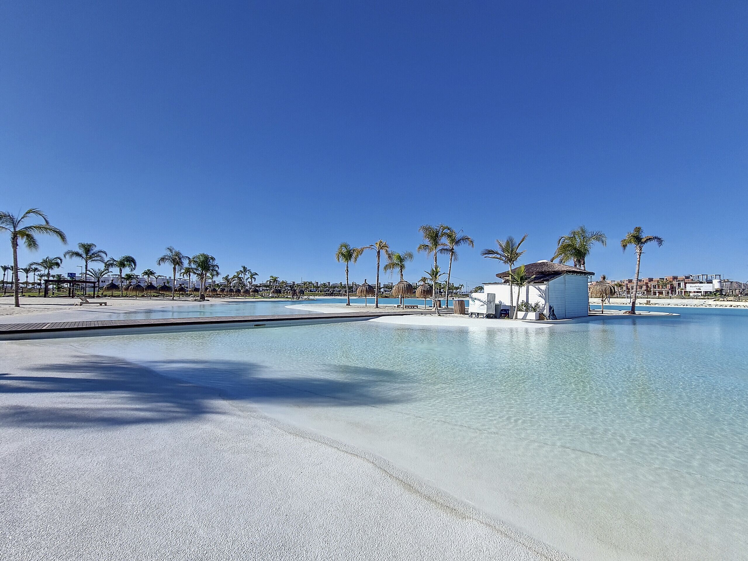 Área de piscina de resort com água cristalina, palmeiras e uma praia arenosa.