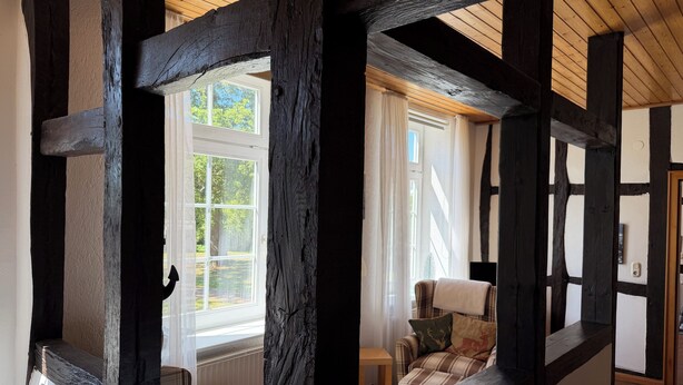 A room with exposed wooden beams, a window with a view of greenery, a plaid sofa, and a small table. - "Sonneneck" - Garden view, half-timbered, 37 m², sleeps 2, dogs welcome (Bösdorf)