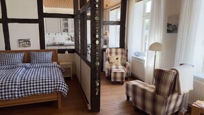 A room with a bed, a chair, a table, a window, and a kitchen area. - "Sonneneck" - Garden view, half-timbered, 37 m², sleeps 2, dogs welcome (Bösdorf)