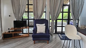 Una sala de estar con un sillón azul oscuro, una silla blanca, un mueble de televisión de madera y ventanas grandes con cortinas.