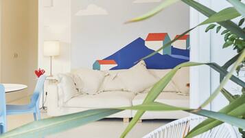A room with a white sofa, a blue chair, a lamp, and a wall with a mountain and house illustration.