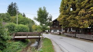 En træbro over en å, en bygning med balkon, og en vej langs en græsplæne.