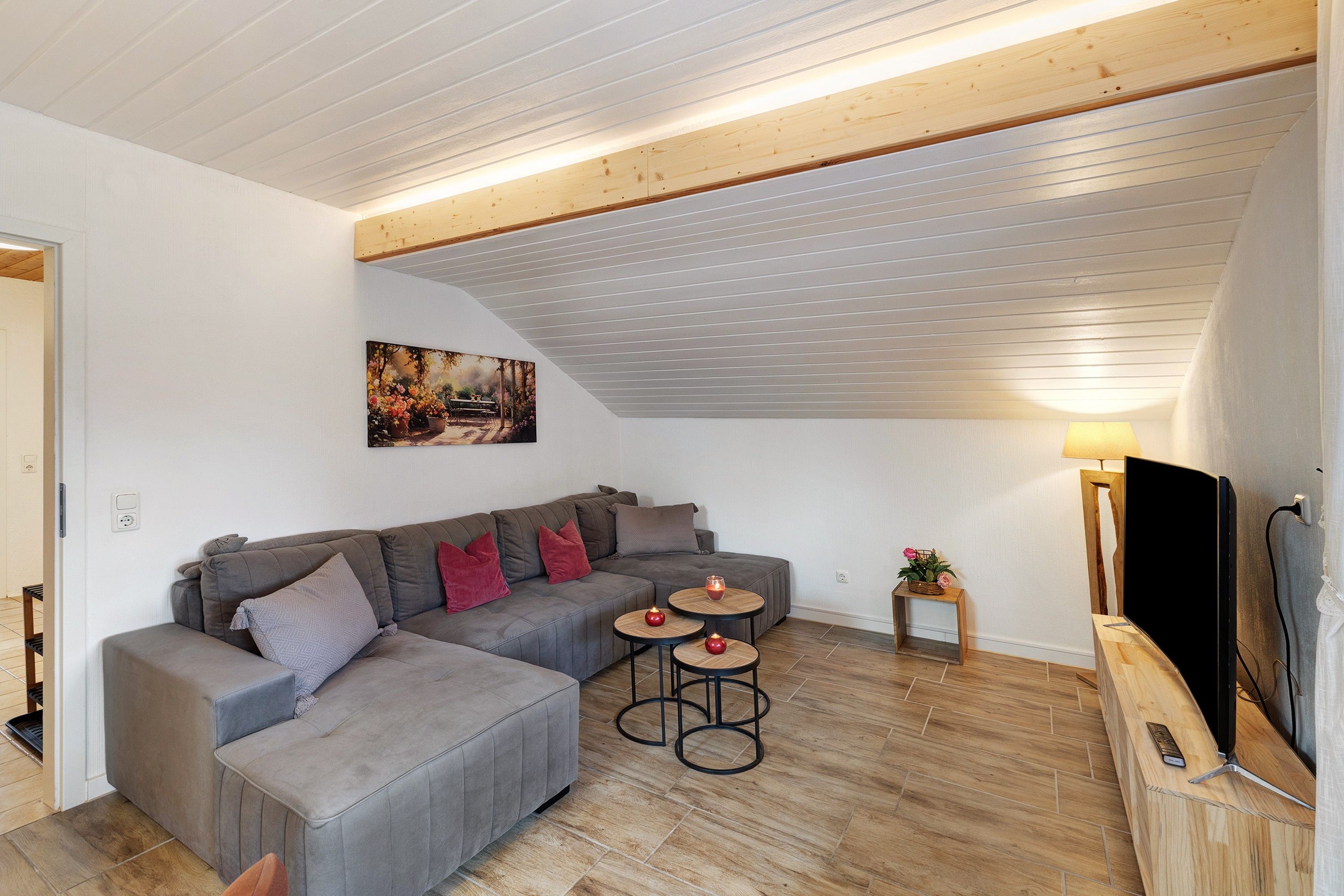 Ein modernes Wohnzimmer mit einem grauen Ecksofa, zwei kleinen runden Tischen, einem Gemälde an der Wand und einem Fernseher auf einem Holzgestell.