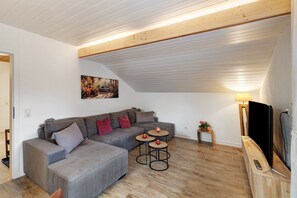 Un salon moderne avec un canapé d’angle gris, deux petites tables rondes, un tableau accroché au mur et un téléviseur posé sur un meuble en bois.