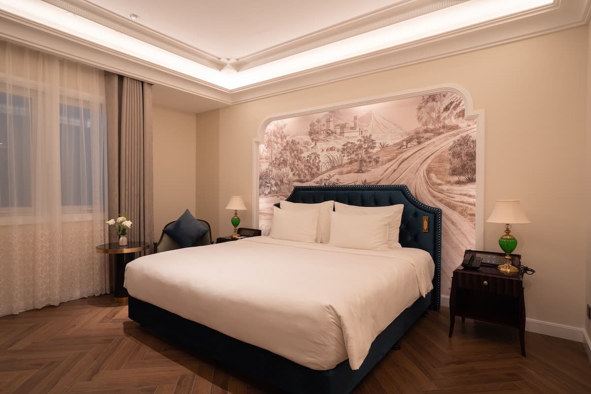 Habitación de hotel con una cama grande, cabecera con un mural de paisaje, mesita de noche con lámpara y ventana con cortinas translúcidas.