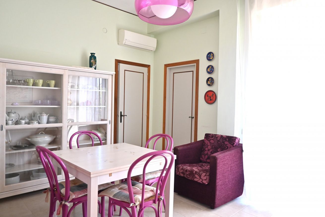 Un coin repas avec une table et des chaises blanches, un meuble avec des portes en verre et un fauteuil violet.