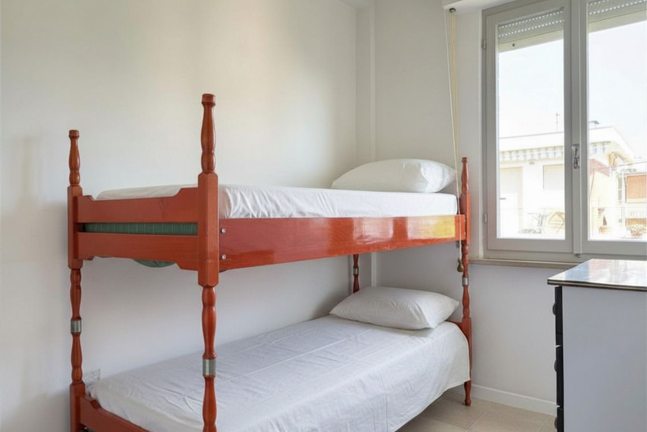 A bunk bed with a lower bunk, a single bed on top, and a window with a view of buildings.