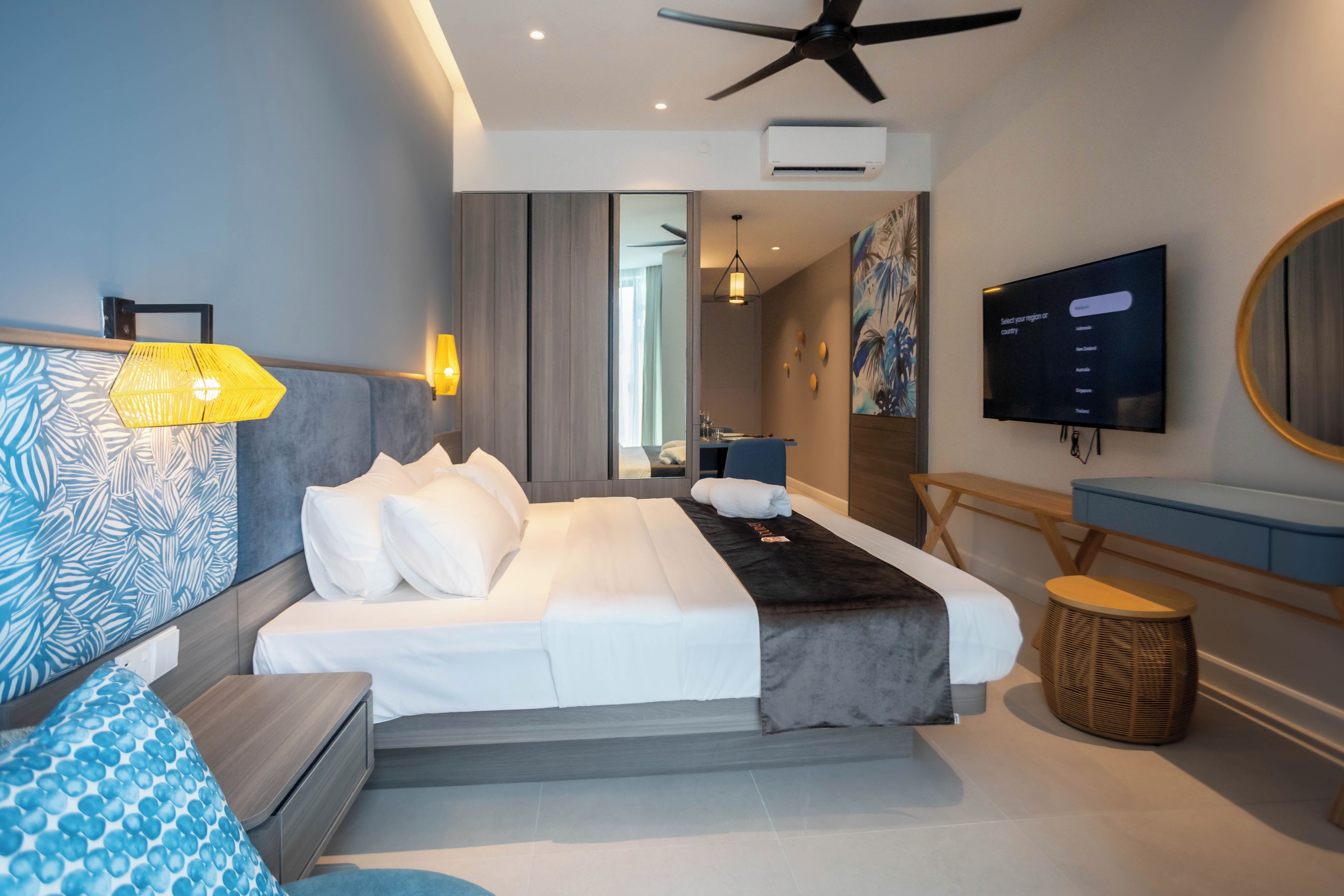 Une chambre d’hôtel moderne équipée d’un lit, d’une table de chevet, d’une lampe, d’une télévision et d’un ventilateur de plafond.