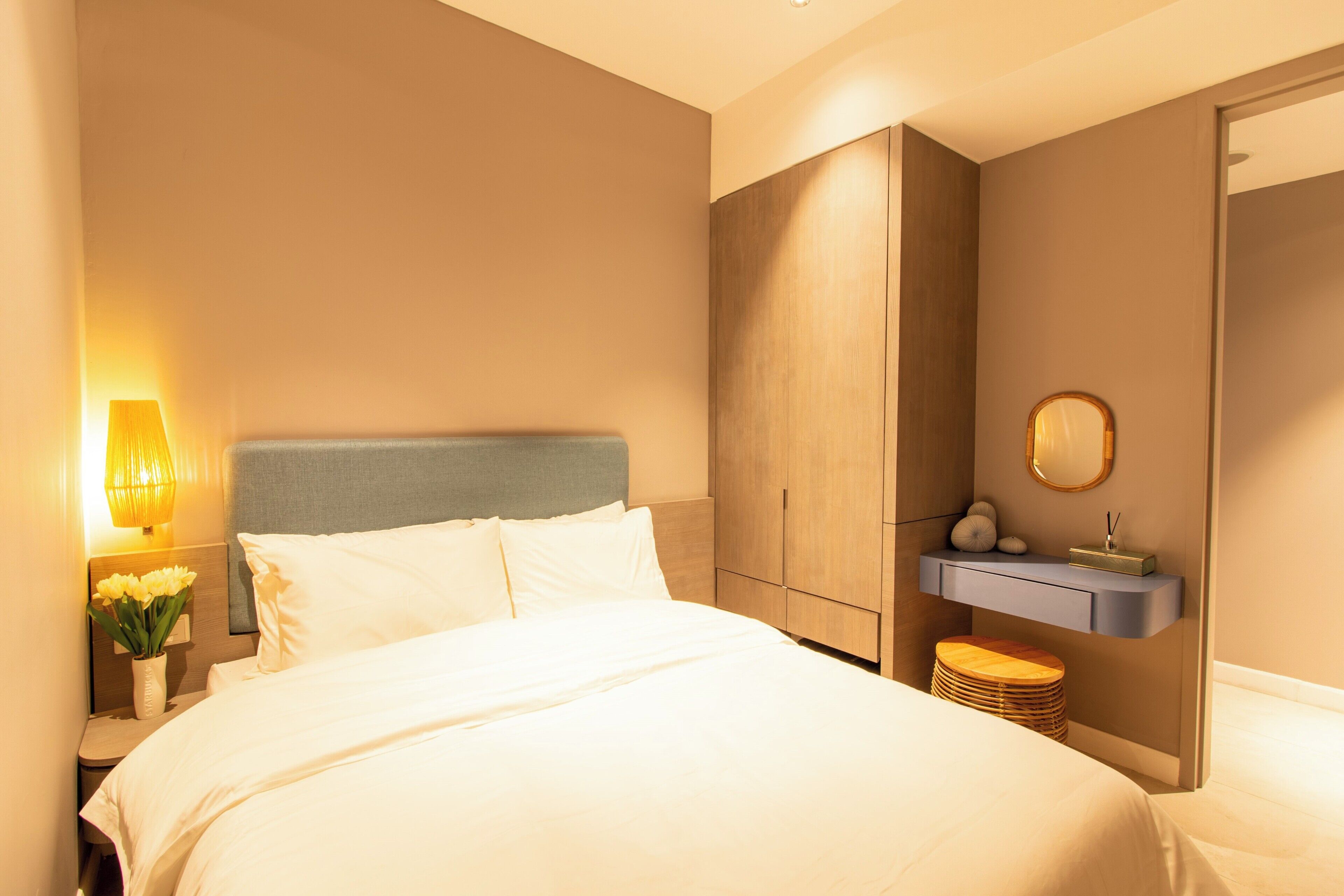 A hotel room with a bed, bedside lamp, nightstand, and a small wooden stool.