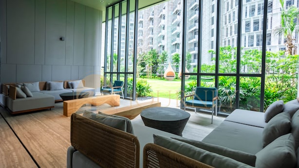 A modern living room with a sofa, chairs, a coffee table, and a view of a garden and buildings through large windows.