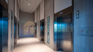 A modern hotel corridor with elevators, a tiled floor, and a glass wall with a logo.