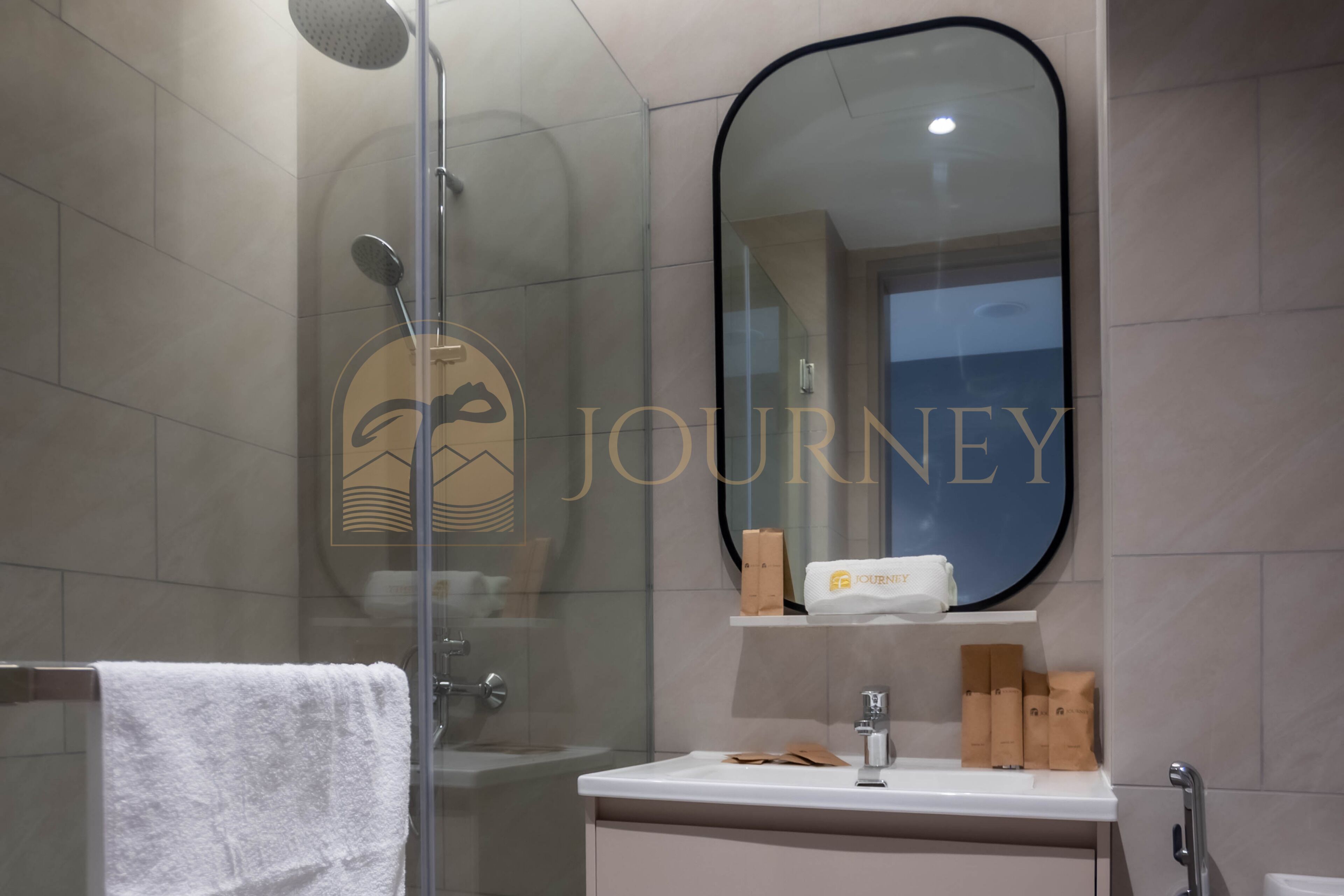 A modern bathroom with a glass shower enclosure, a white sink, and a mirror with a logo.