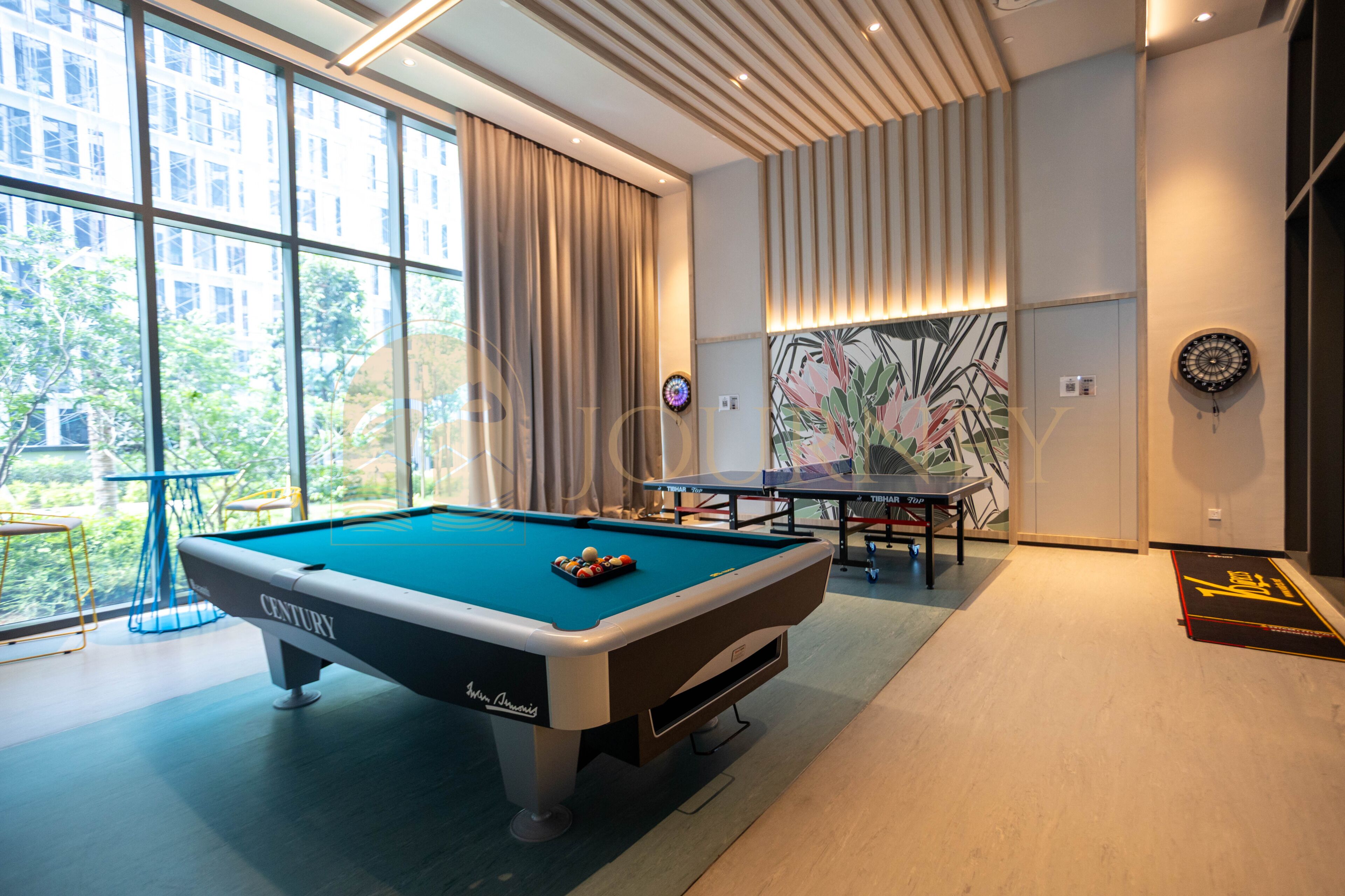 A pool table with a billiard cue, a ping pong table, and a dartboard in a room with large windows and a view of buildings outside.