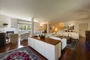 Een ruime woonkamer met een centraal eiland, witte banken, een eethoek met stoelen en een trap naar een hoger niveau.