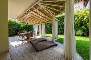 A covered outdoor area with a table and chairs, a lounge chair, and a garden with greenery. - Villa Camillo by MMega (Forte dei Marmi)
