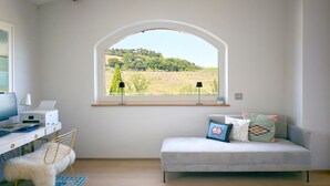 Una stanza con una scrivania, una sedia, un divano, una finestra vista verde e una lampada.