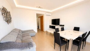 Un salon moderne comprenant un canapé d’angle, un coin repas avec une table et des chaises, un téléviseur à écran plat et un appareil de climatisation.