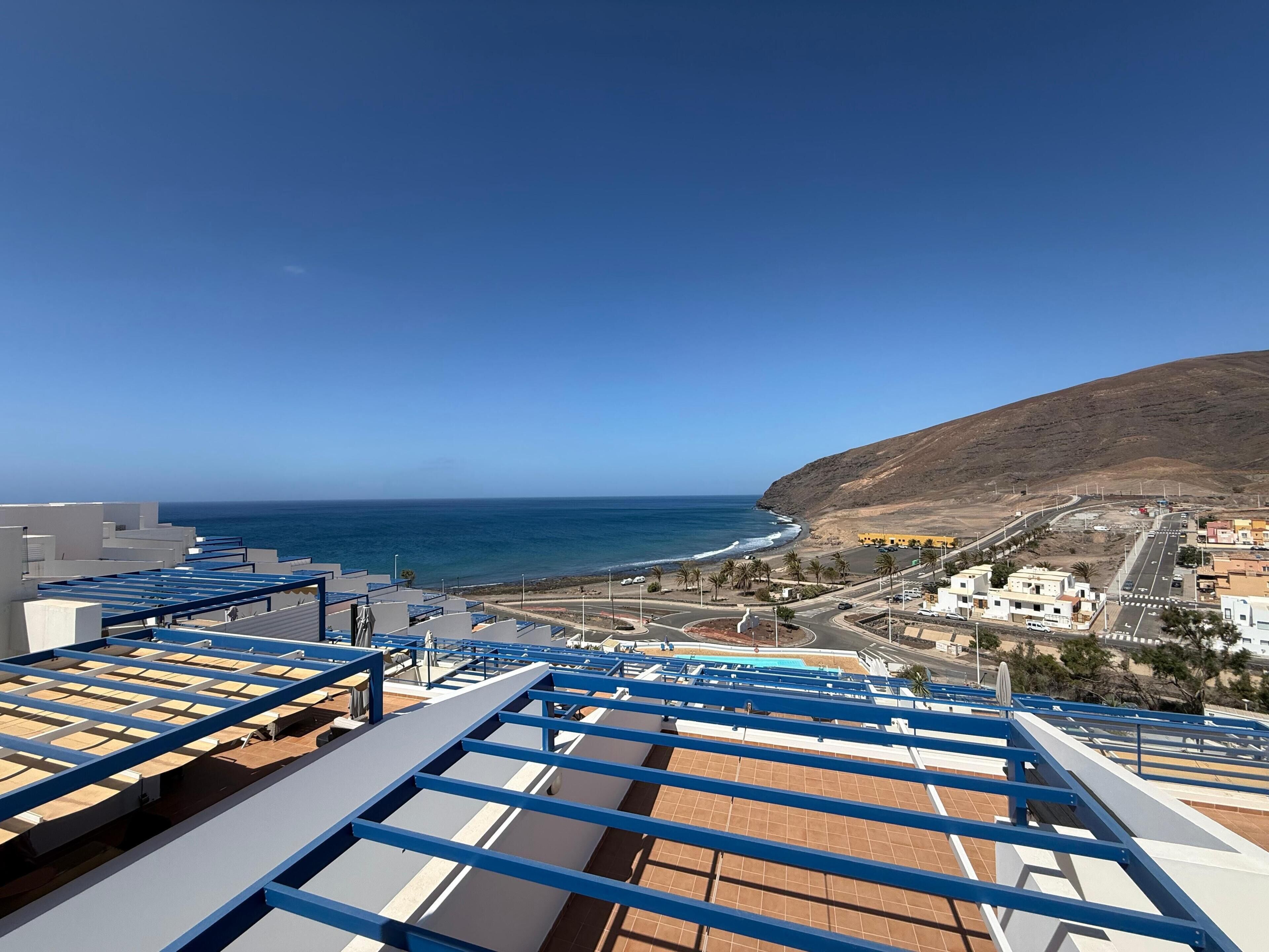 A coastal view with a clear sky, a beach, and a hilly landscape.