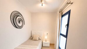 Une petite chambre à coucher, bien rangée, avec un lit simple, une table de chevet avec une lampe, une œuvre d’art accrochée au mur et une fenêtre avec des rideaux.