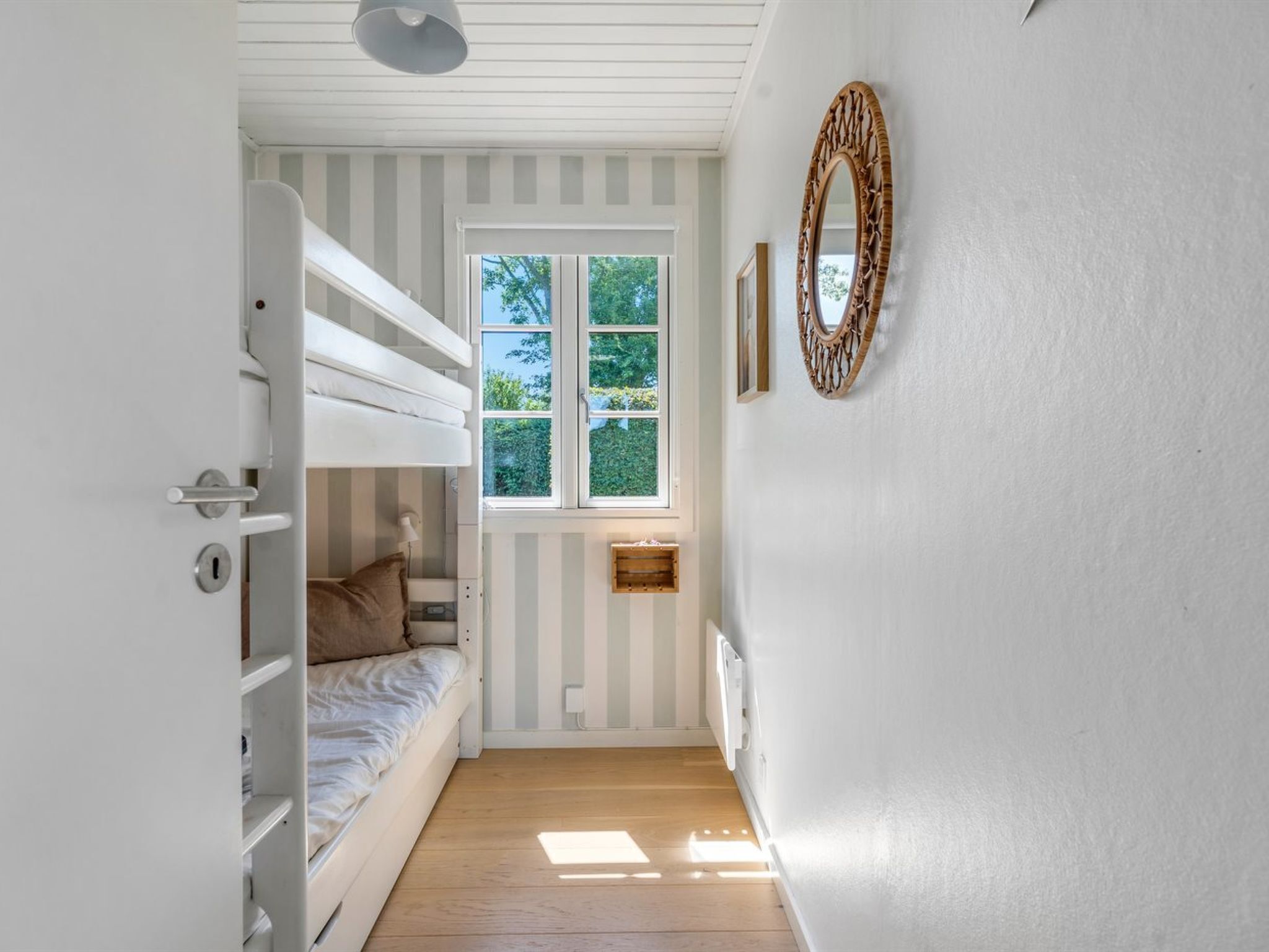 A room with a bunk bed, a window with a view of greenery, a wall-mounted mirror, and a small wooden shelf.