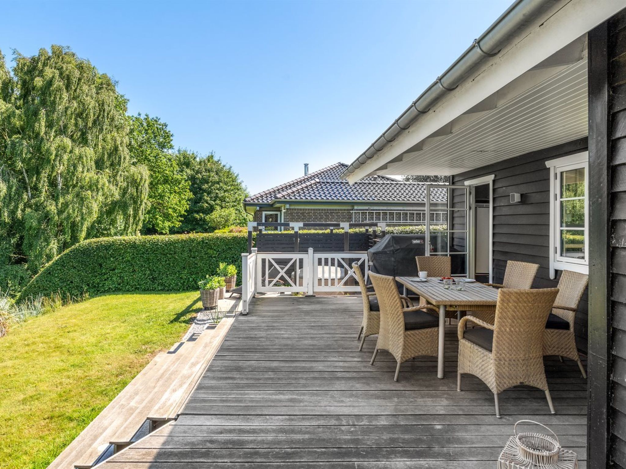Een houten terras met buitenmeubilair, een barbecue en uitzicht op een huis met een pannendak.