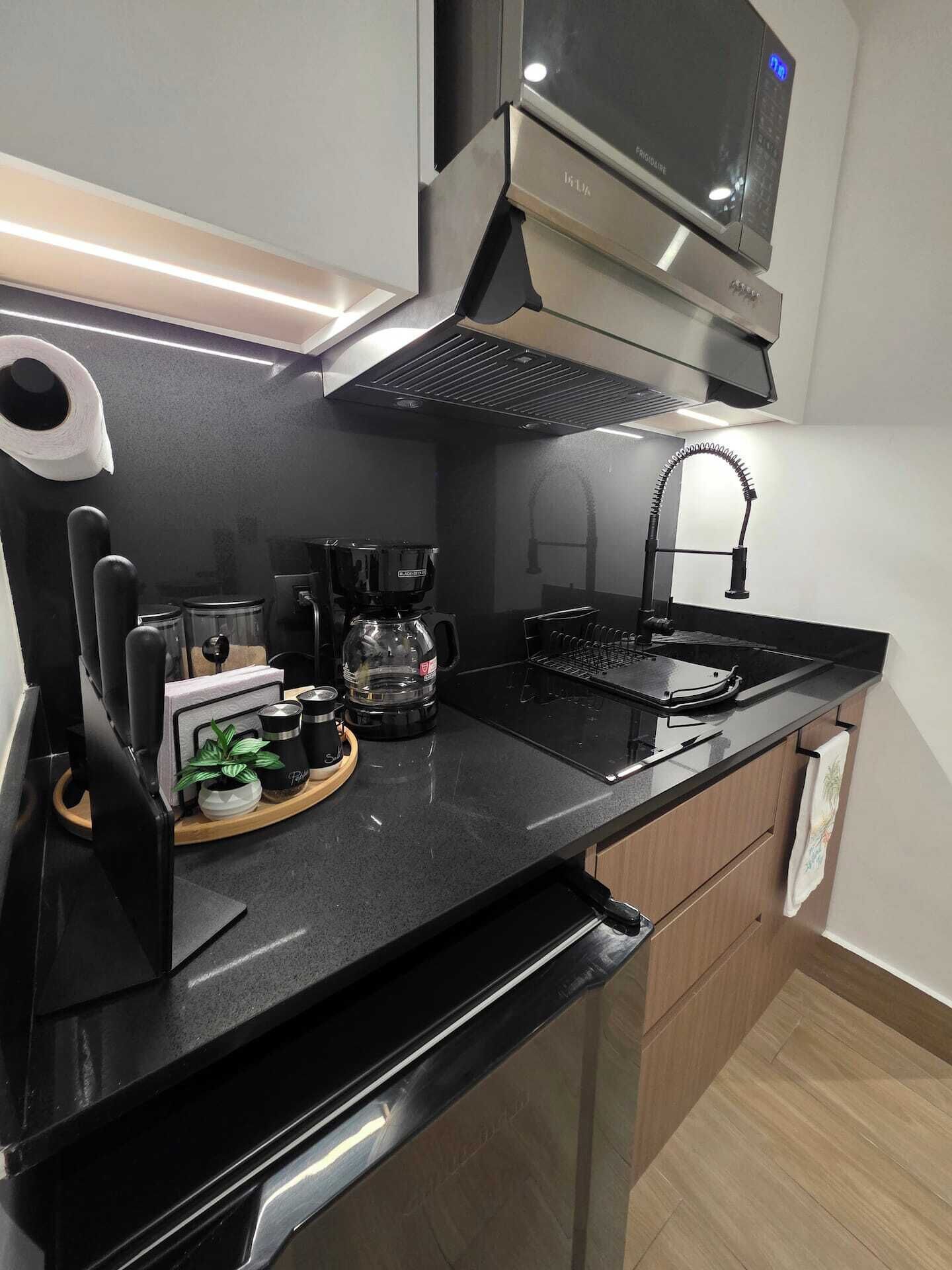 Uma cozinha moderna com bancada preta, eletrodomésticos de aço inoxidável, cafeteira e micro-ondas.