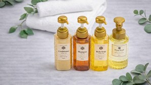 Quatre flacons de produits de soins personnels sont posés sur une surface, avec une serviette blanche et des feuilles vertes en arrière-plan.