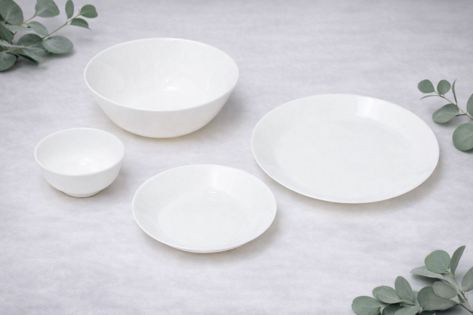 Four white ceramic bowls of varying sizes on a marble surface with eucalyptus leaves.