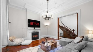 A living room with a fireplace, a sofa set, a TV displaying 'NETFLIX,' and a chandelier. - Walk to Allentown With Free Parking WFH Space (Buffalo)