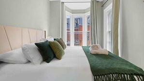 A hotel room with a bed, pillows, a green throw blanket, and a view through a window. - Walk to Allentown With Free Parking WFH Space (Buffalo)