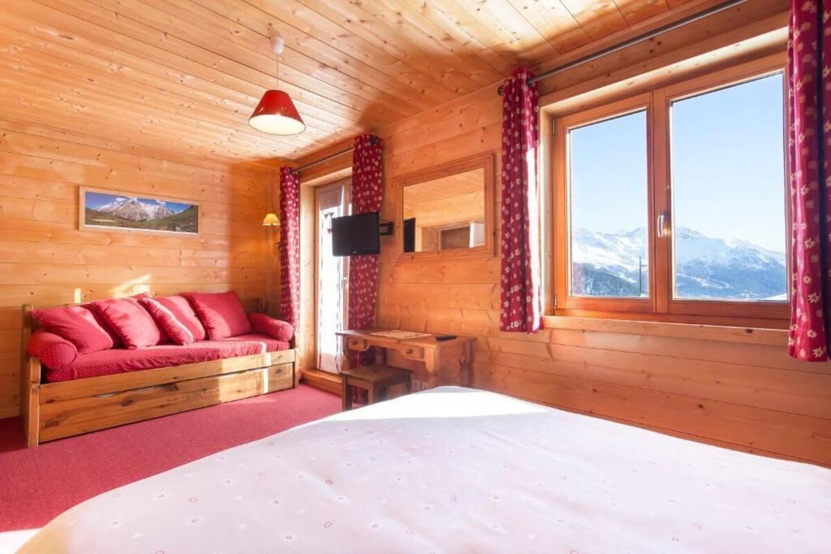 A wooden cabin room with a bed, a wooden desk, a television, and a window with a mountain view.