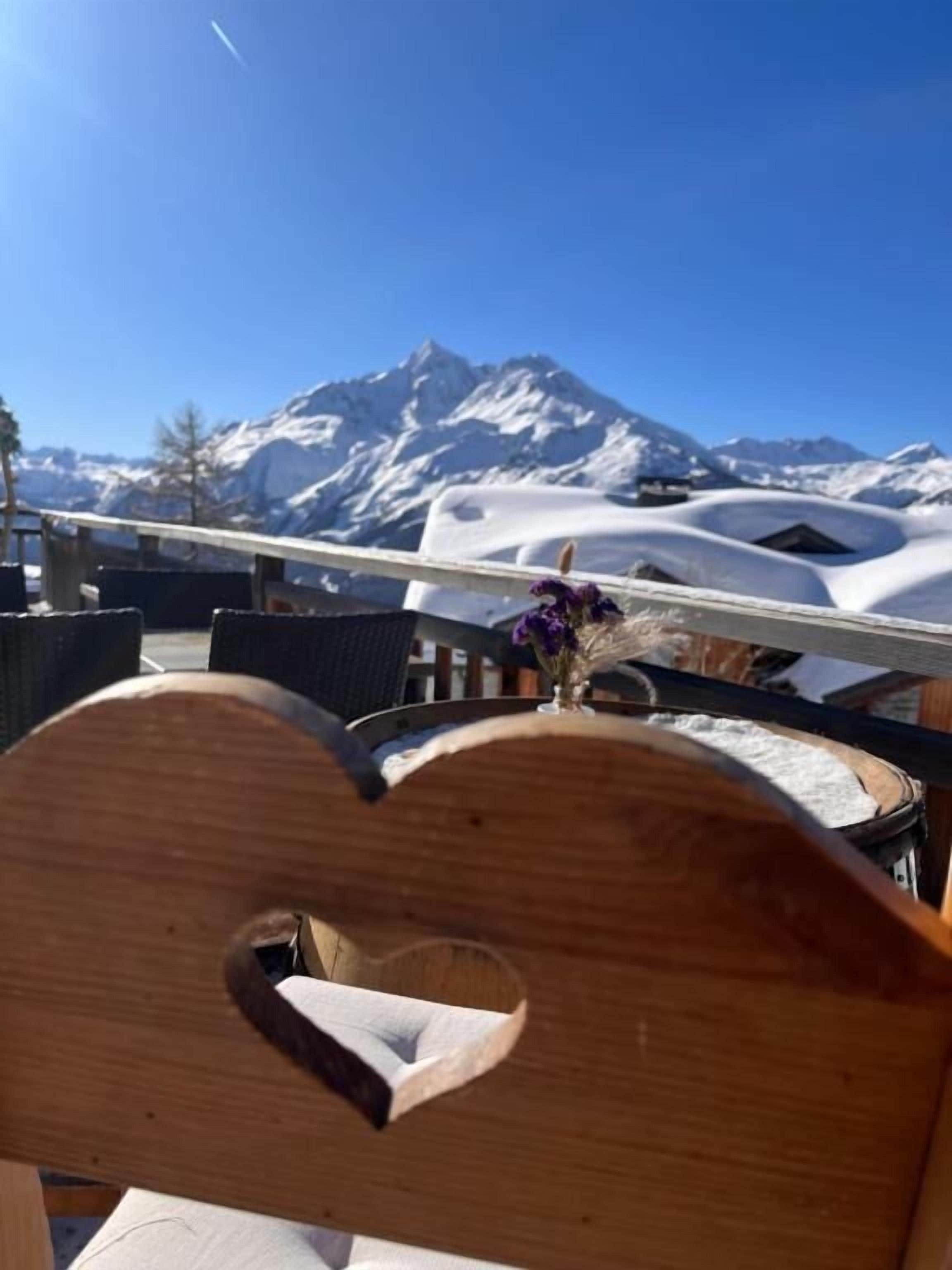 A wooden chair with a heart-shaped cutout, a window with a view of snow-covered mountains, and a clear blue sky.