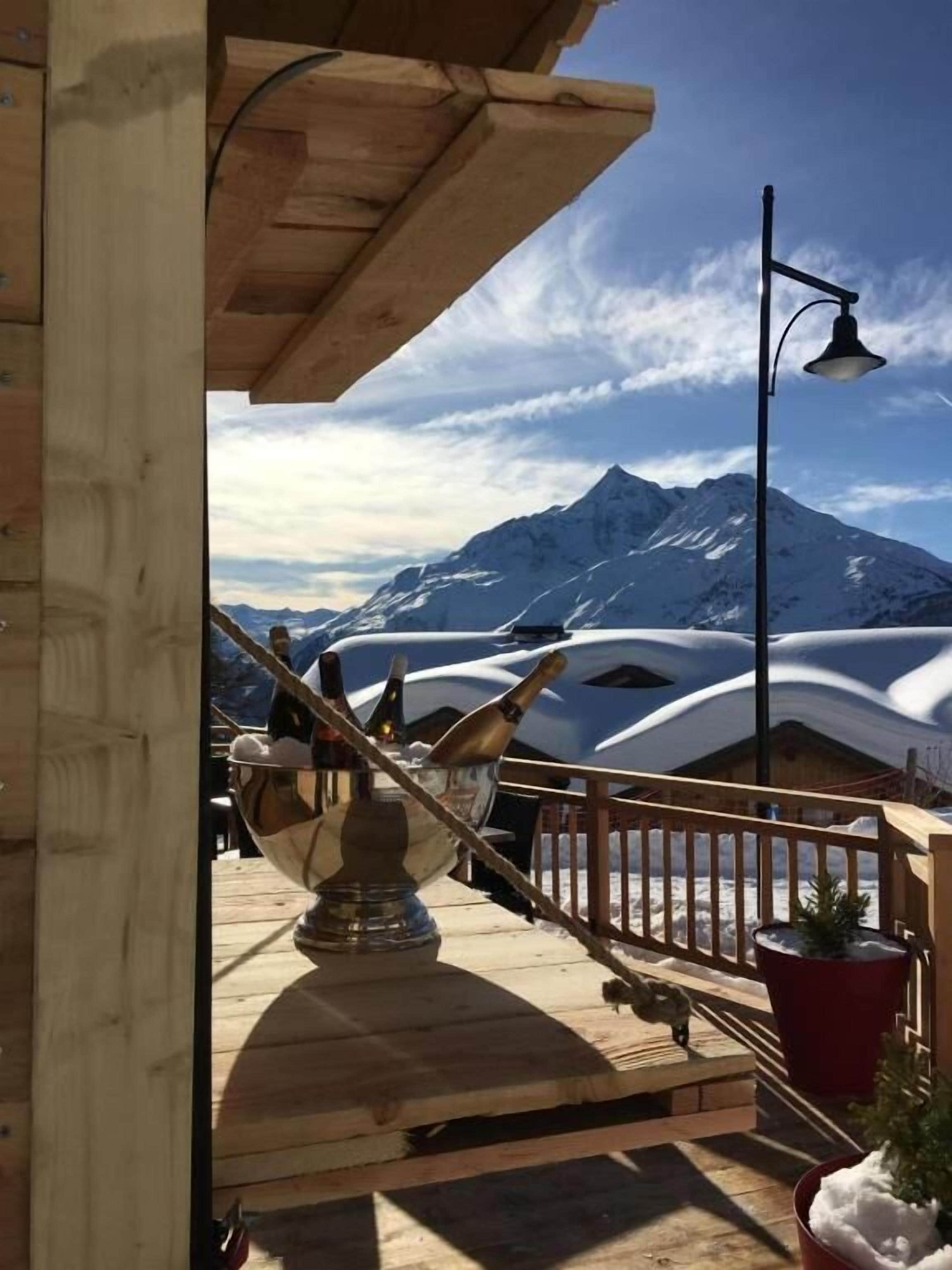 Une terrasse en bois avec un récipient en métal et une cloche, une plante en pot rouge et une vue sur la montagne.