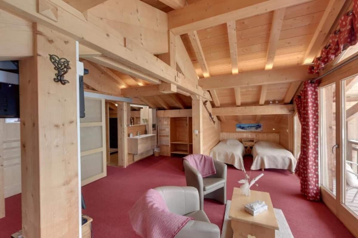 A wooden interior with a high ceiling, a bed, a chair, a small table, and a window with curtains.