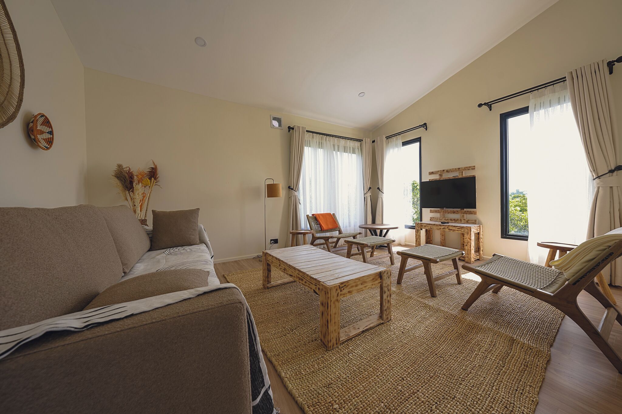 Una sala de estar con sofá, mesa de centro, sillas, televisor y ventanas amplias con cortinas.