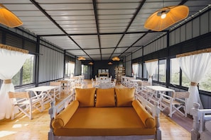 Amplio espacio interior con mesas y sillas dispuestas para comer o conversar, ventanas grandes a los lados y una zona central con cojines.