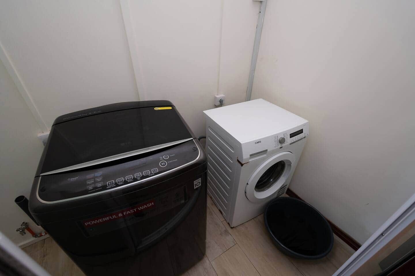 Ein kleiner Waschraum mit einer schwarzen Waschmaschine und einem Trockner, einem weißen Trockner und einer schwarzen Mülltonne.