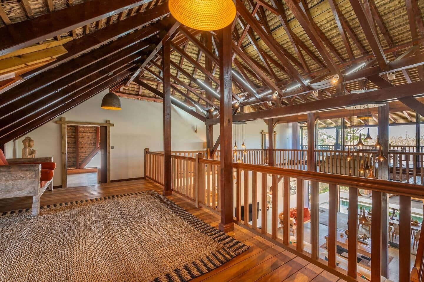 Un ático con vigas de madera, una escalera, una lámpara colgante y vistas al exterior.