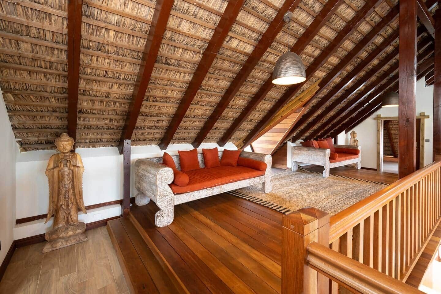 Quarto em estilo loft com teto inclinado, vigas de madeira, escada de madeira, um sofá e uma estátua.