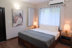 A bedroom with a bed, a nightstand, a wall-mounted air conditioner, a painting, and a window with curtains. - Luxe 2BHK Private Garden Flat by ORR Tech Parks (Bengaluru)