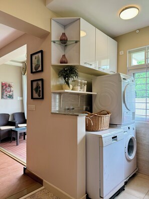 A laundry area with a washing machine, shelves with items, a wicker basket, and a potted plant. - Luxe 2BHK Private Garden Flat by ORR Tech Parks (Bengaluru)