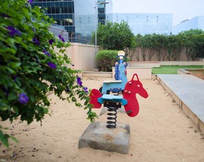 A playground with a seesaw and a climbing structure, surrounded by greenery and modern buildings in the background. - Luxe 2BHK Private Garden Flat by ORR Tech Parks (Bengaluru)