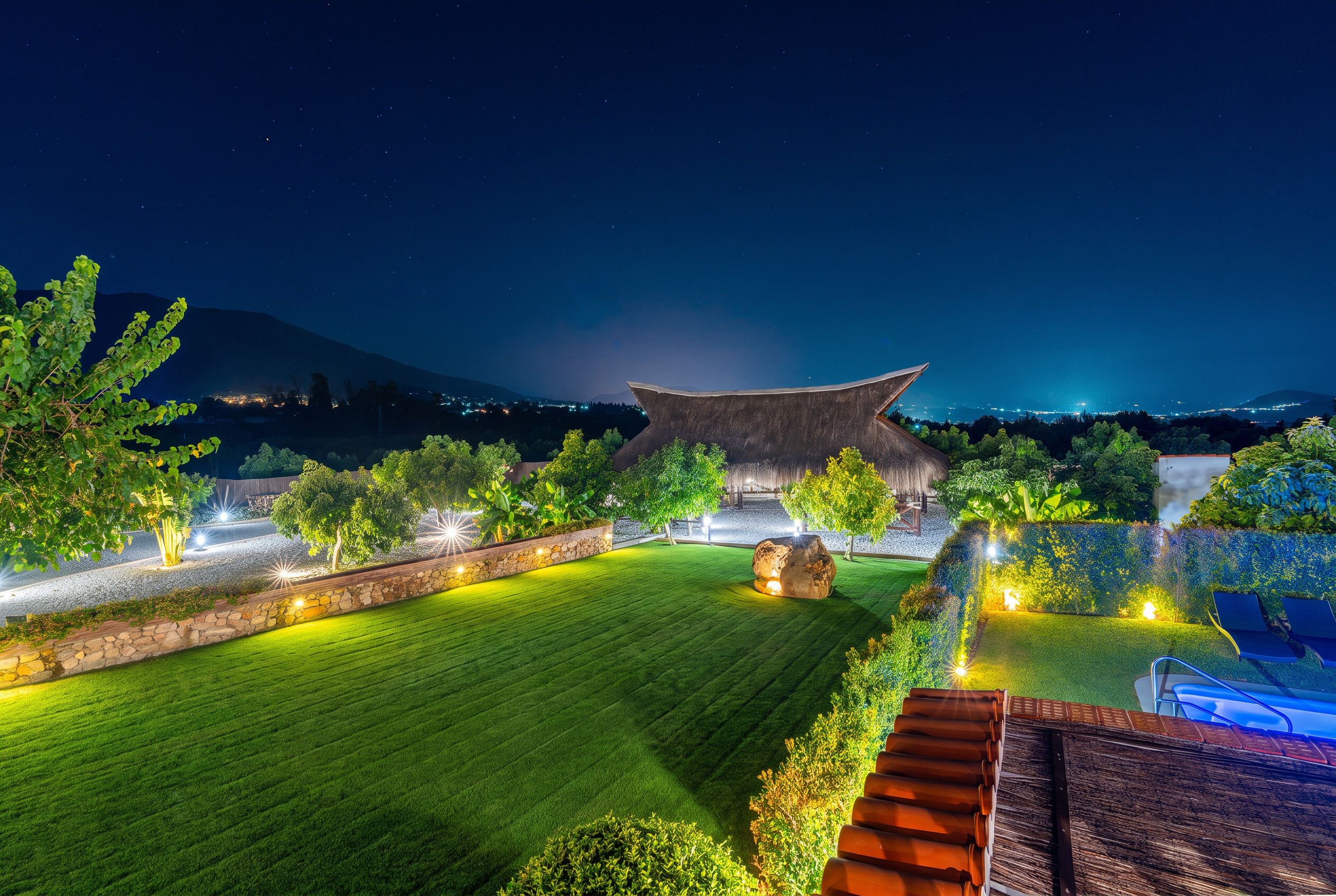 A well-lit garden with a traditional roof structure, a stone wall, and a pool area.
